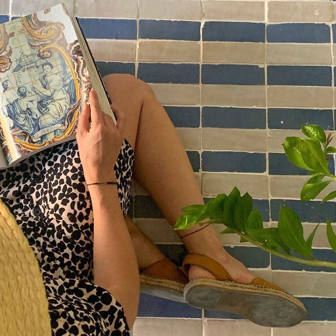 Person sitting on a blue and white tiled floor holding a book, wearing sandals and a patterned outfit.