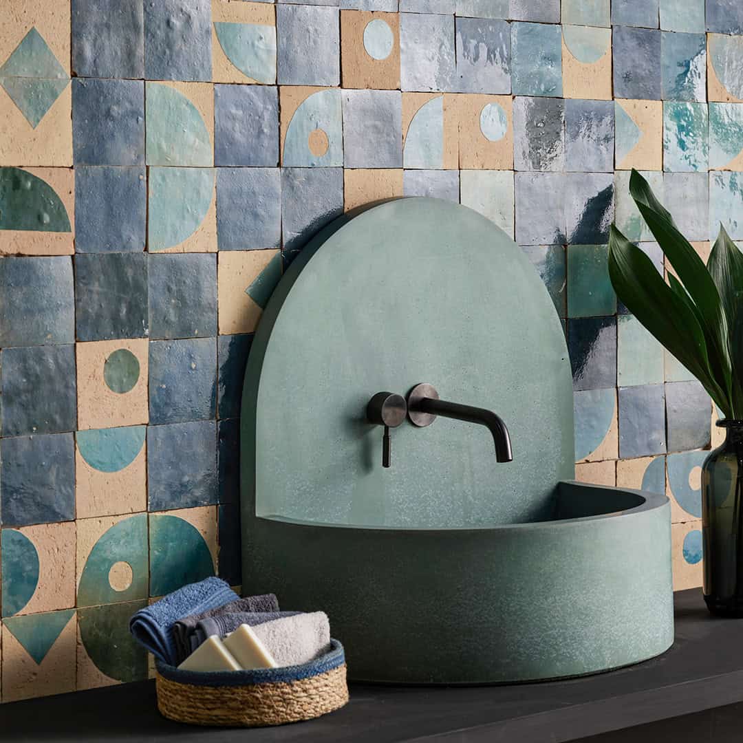 Green ceramic sink against a patterned tile wall with a plant and soap on a shelf.