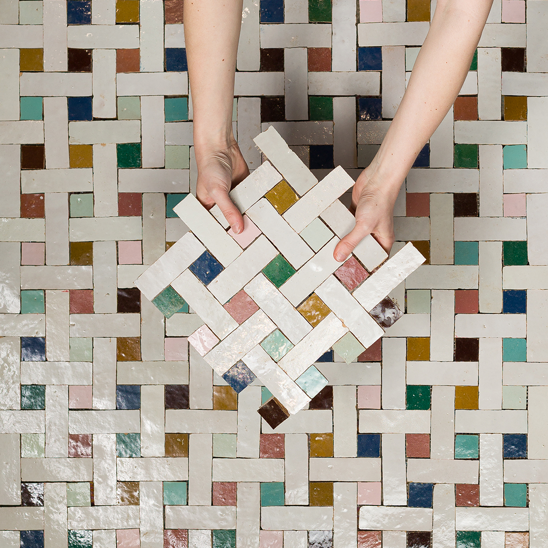 Hands holding a woven tile sample against a multicolored tile background
