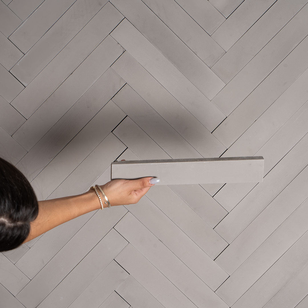 A rectangular herringbone pattern in light gray with a smooth matte finish, ideal for modern, minimalist, or industrial interior flooring and wall design applications.
