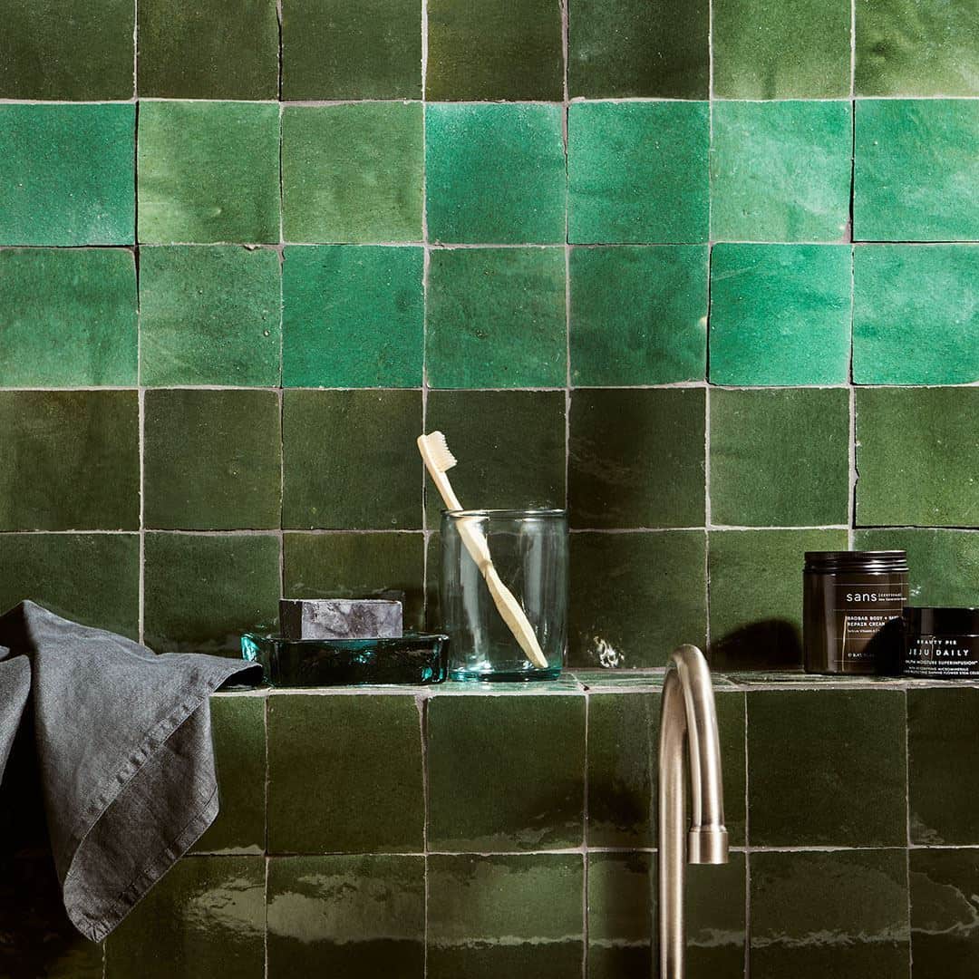 Green tiled bathroom wall with a glass, toothbrush, and toiletries.