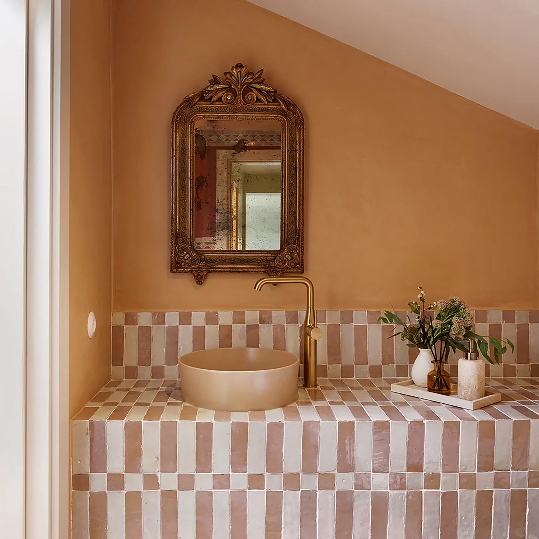 Elegant bathroom with terracotta walls, beige vessel sink, gold faucet, ornate mirror, and decorative tray featuring flowers and candle beside tiled beige and pink backsplash.