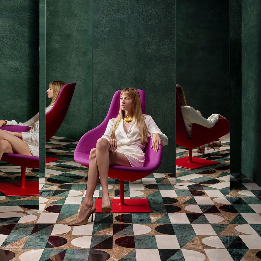 Woman sitting on a purple chair in a room with mirrors and geometric floor patterns.