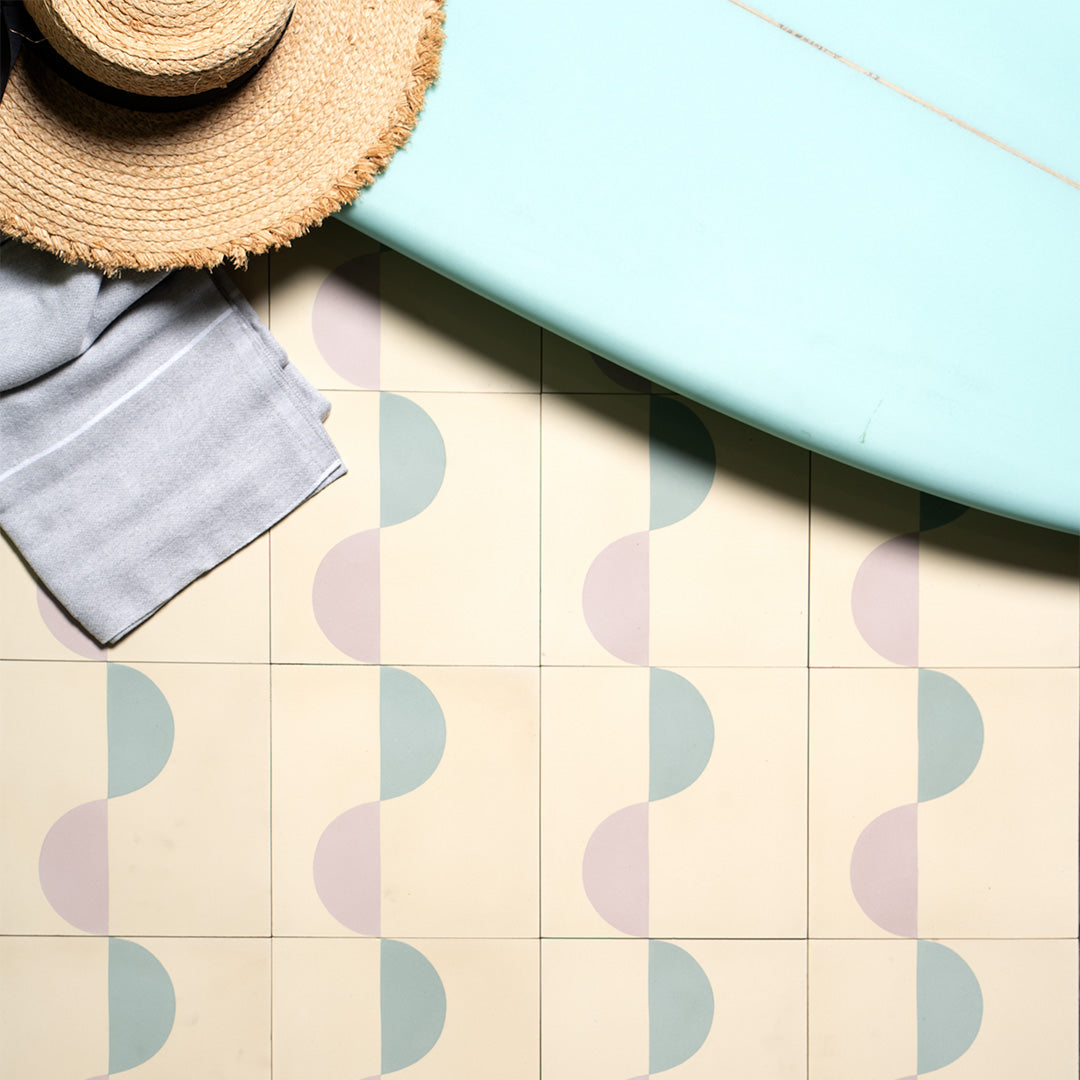 Surfside-inspired cement tiles featuring pastel blue and mauve wave patterns on a cream background, styled with a straw hat, gray towel, and light blue surfboard for a coastal aesthetic.