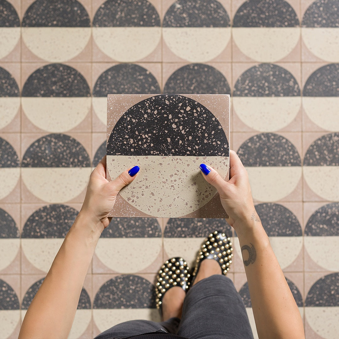 A square cement tile featuring a large circle divided into black and beige halves with terrazzo speckles, set against a warm brown background for a modern geometric look.
