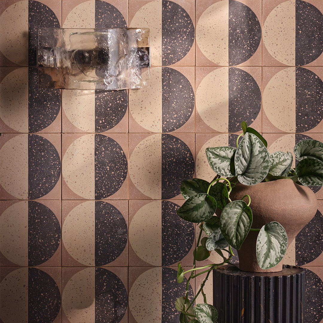 A square cement tile featuring a large circle divided into black and beige halves with terrazzo speckles, set against a warm brown background for a modern geometric look.