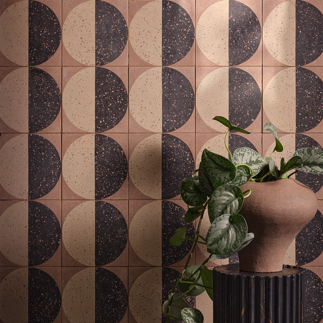 A square cement tile featuring a large circle divided into black and beige halves with terrazzo speckles, set against a warm brown background for a modern geometric look.