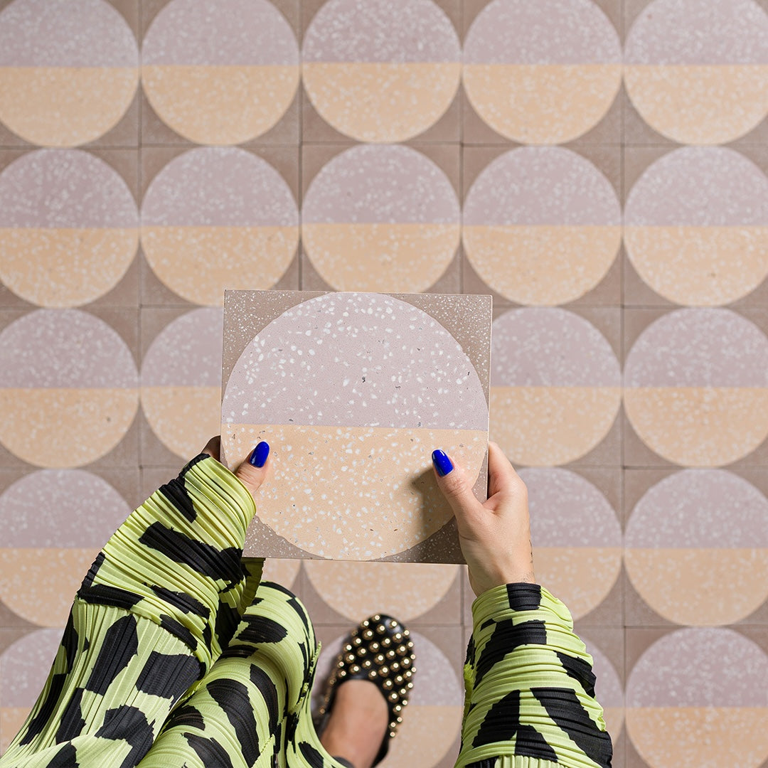 A square cement tile featuring a large circle divided into peach and mauve halves with white terrazzo speckles, set against a taupe background for a modern geometric look.