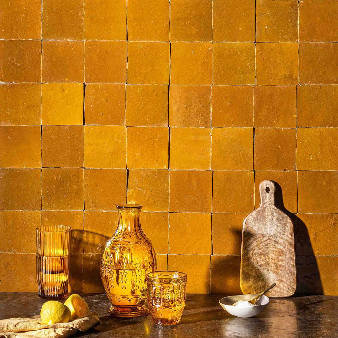 Decorative setting with a bottle, glasses, and cutting board against a yellow tiled wall.