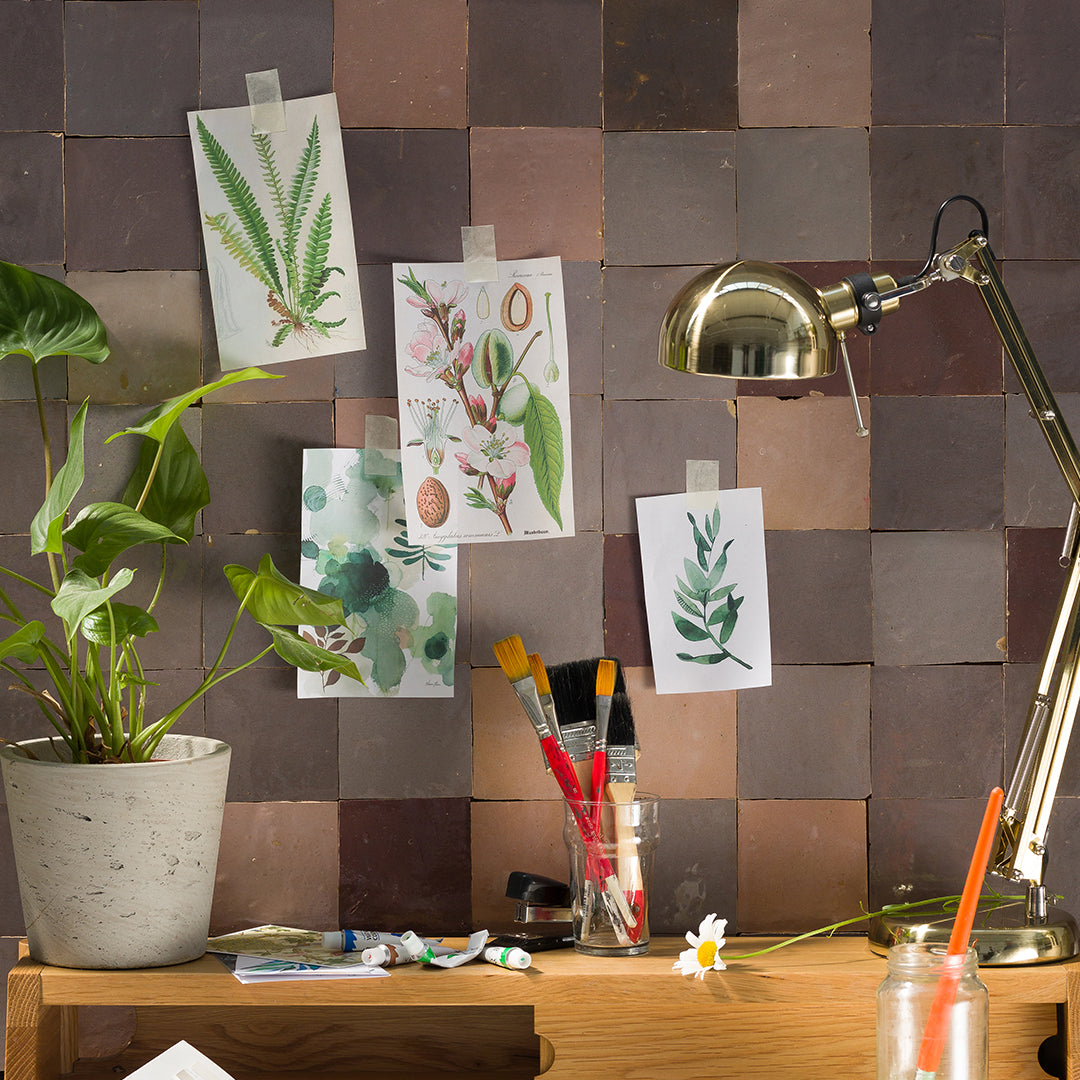 Desk with plants, art supplies, and decorative prints against a tiled wall.