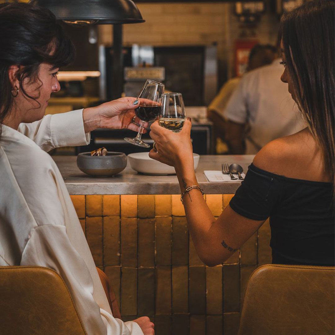Two women at a bar counter clinking wine glasses in a warm, dimly lit setting.