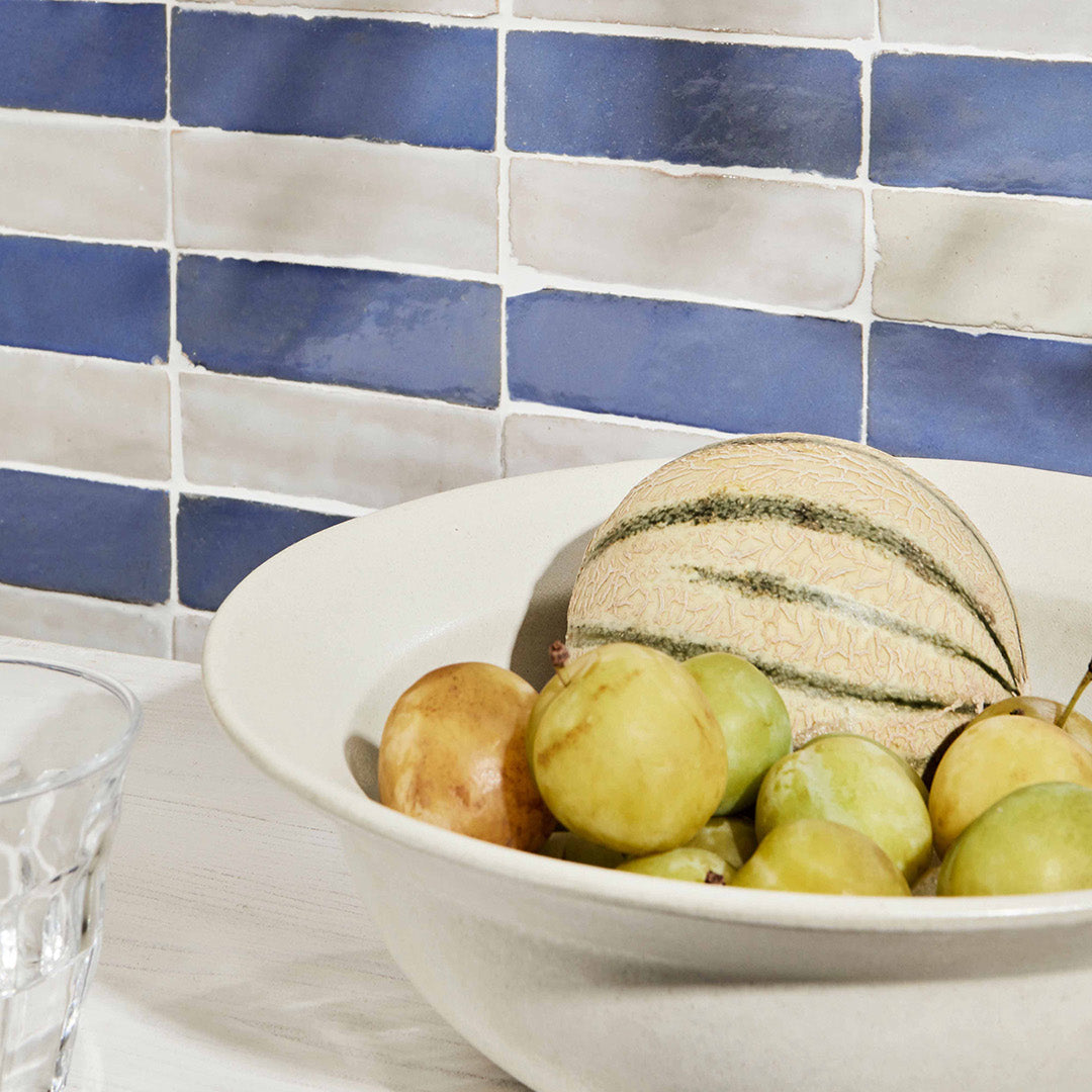 Bowl of fruit including apples and a melon on a table with a blue and white tiled wall in the background.