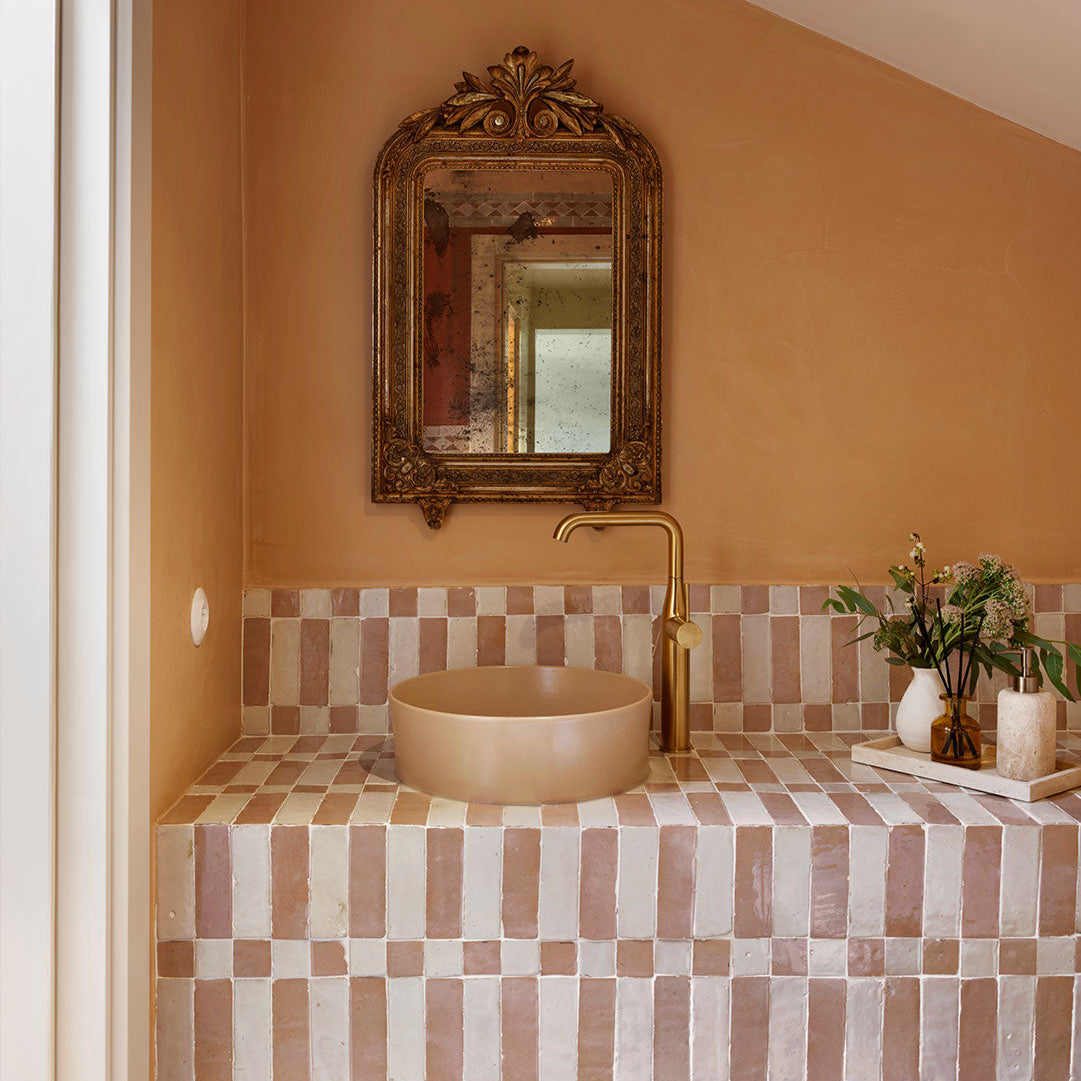 Bathroom with beige sink, gold faucet, and decorative mirror on a checkered tile wall.