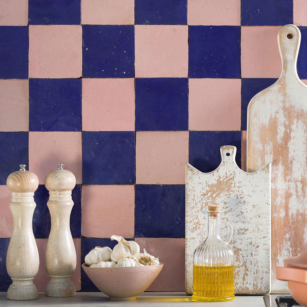 Pink and dark blue checkerboard kitchen wall tiles with rustic cutting boards, garlic bowl, pepper mills, and cooking oil.