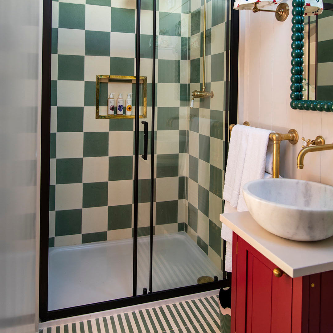 Modern bathroom with a glass shower enclosure featuring green and pure white cement tiles in a checkerboard pattern, accented by brass fixtures, red vanity, and marble sink.