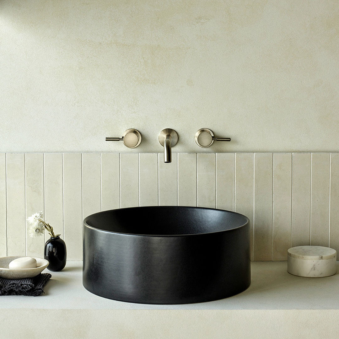 Bathroom interior featuring vertical dirty white cement tiles behind a matte black round basin, brushed nickel wall-mounted faucet, and minimalist decor with neutral, modern design aesthetics.