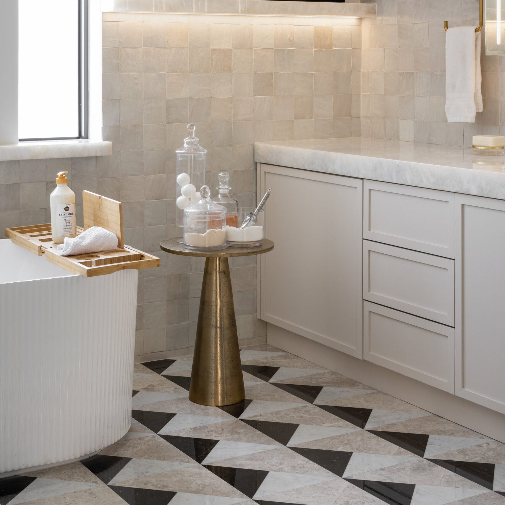 A modern bathroom interior with white cabinetry, a freestanding tub, a small gold side table, patterned black-and-white tile flooring, and a framed black-and-white wall art piece with a gold abstract eye design above the vanity.