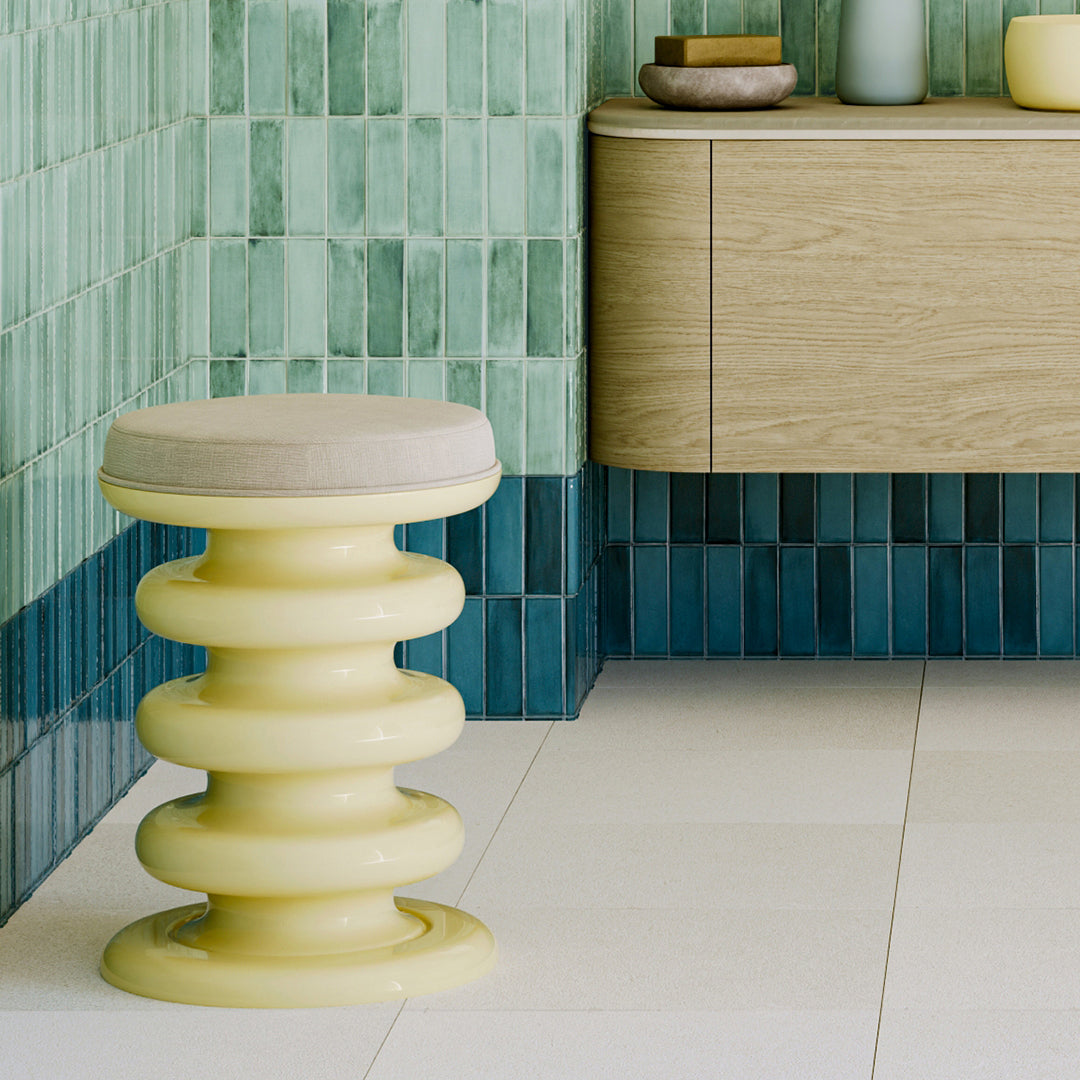 Yellow stool with a cushioned seat in front of a tiled wall and wooden cabinet.