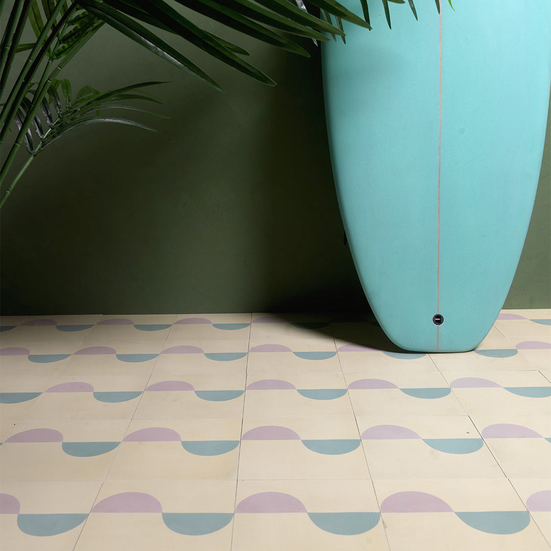 Surfside-inspired cement tiles with pastel blue and mauve wave patterns on a cream base, paired with a teal surfboard and green wall, evoking a coastal, tropical vibe.